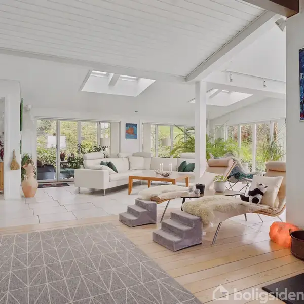 Et lyst stueområde med hvide vægge og loft, hvor to moderne stole og et sofabord er placeret. Store vinduer slipper naturligt lys ind, og planter pynter rummet. En farverig kunstværk hænger på væggen.