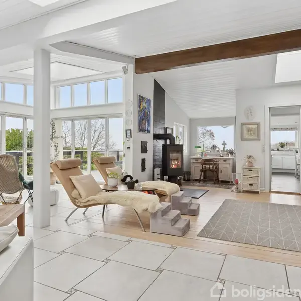 Stue med moderne indretning; lænestole står omkring et lavt bord nær store vinduer. I baggrunden er en brændeovn og åbent køkken. Lyse farver og kunst på væggene.