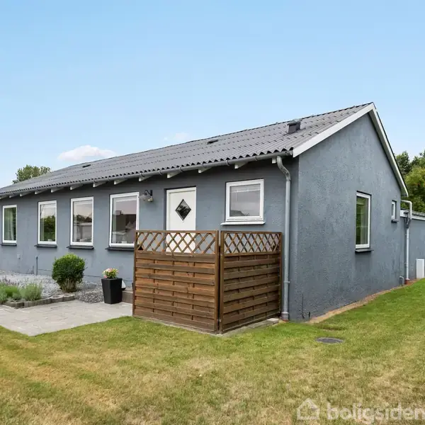Gråt hus med hvidt tag, placeret i en grøn have med græsplæne og hæk. Huset har vinduer på siden og en terrasse med trægitter.