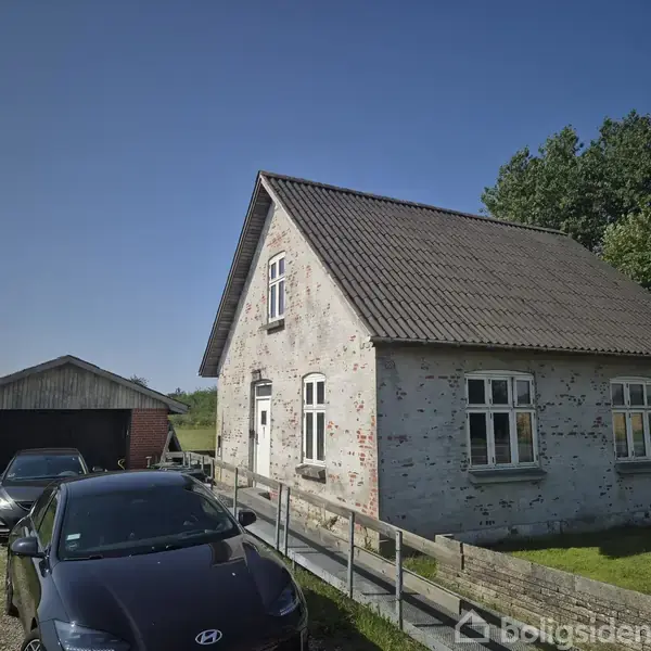 Et ældre, hvidt murstenshus med skråt tag står ved en grusvej. To biler er parkeret ved siden af huset, og en lade ses i baggrunden, omgivet af grønt landskab.