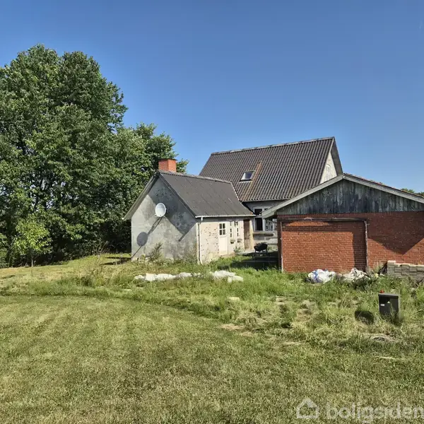 Et landhus står stille i en grøn græsplæne omgivet af træer. Huset har en brun tag, hvid facade, og en murstensvæg til venstre. En satellitskål er installeret.