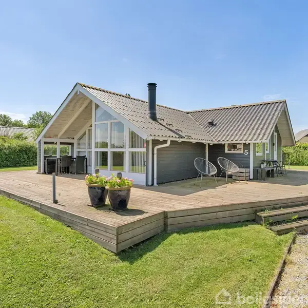 Et moderne sommerhus står på en stor træterrasse. Huset har store glaspartier og en skorsten. Haven er velholdt med grøn græsplæne og buske i baggrunden.