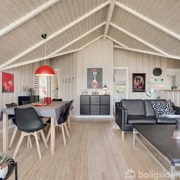 Trævægge og loft i stue med sorte lædersofaer omkring et sofabord. Spisebord til venstre med rødt pendel, kunst på væggene og køkken udsyn i baggrunden.