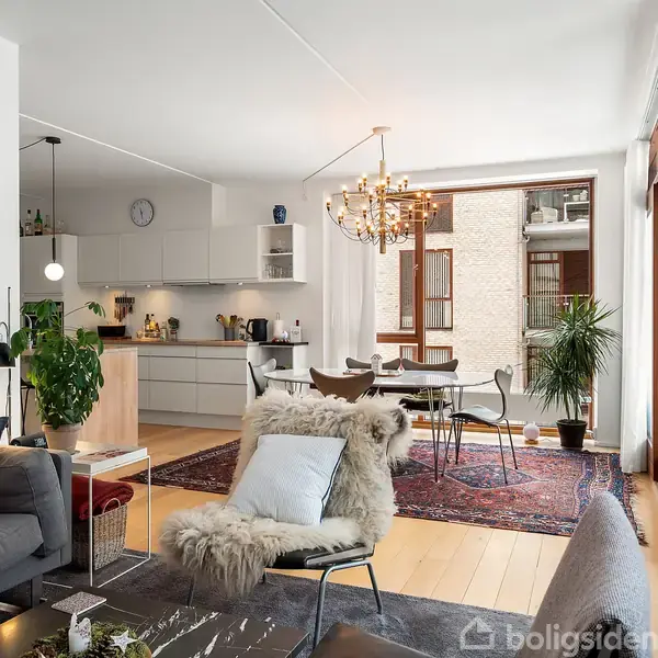 Sofa med puder står i en moderne opholdsstue, udsmykket med en stor lysekrone over et spisebord. Rummet har trægulve, planter og store vinduer mod en altan.