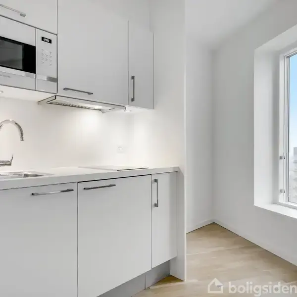 Et funktionelt, moderne køkken med hvide skabe, indbygget mikrobølgeovn og stålarmatur. Sollys strømmer ind gennem et stort vindue, der viser byudsigten.