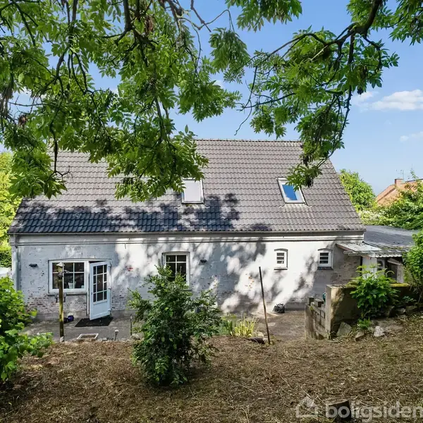 Et hvidt hus med gråt tag står i en frodig have. Der er grønne træer omkring, og en skyfri himmel ses i baggrunden.