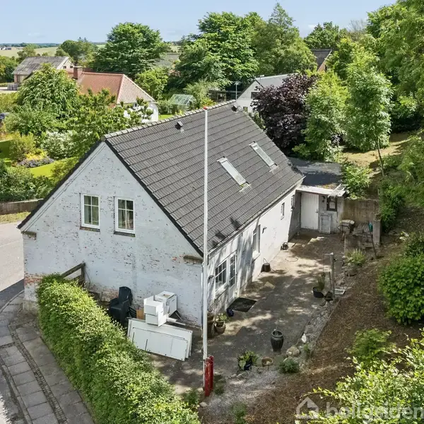 Et hvidt hus med sort tag ligger ved en vejkant. Foran er en lille gårdhave med planter. Området er omgivet af grønne træer og buske.
