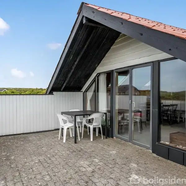 Verandaen har plastikstole og et bord, omgivet af en hvid træbaldakin. Store glasdøre giver adgang til huset, der ligger i et beboelsesområde med omkringliggende huse.