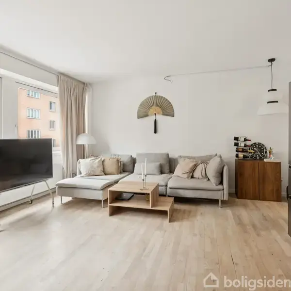Sofa med puder står i stue, flankeret af et bord og en lampe. Et tv står foran et stort vindue. Køkken er delvis synligt med køleskab.