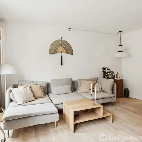 Grå hjørnesofa med puder står i en lys stue. Foran sofaen er et træbord med stearinlys. En dekorativ vifte hænger på væggen, og et køleskab er synligt i højre hjørne.
