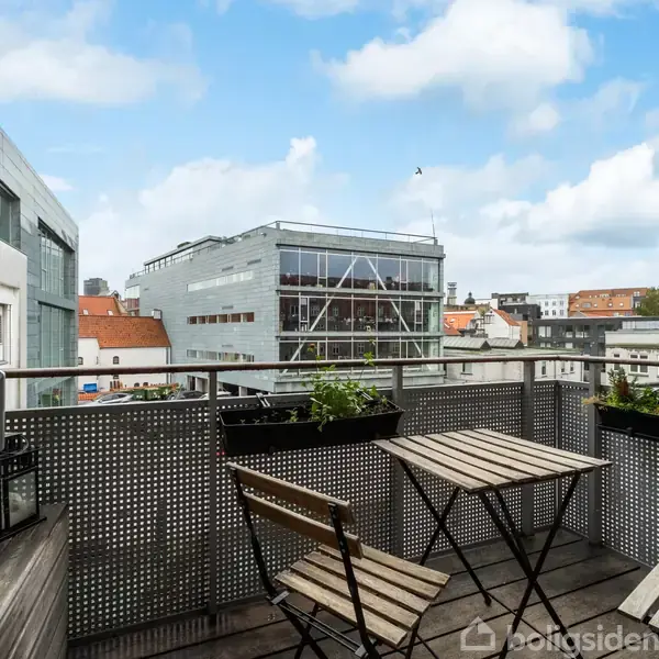 En lille altan med et bord og to stole af træ er omgivet af moderne bygninger og blå himmel. Planteliv i altankasser tilføjer grønt til den urbane udsigt.