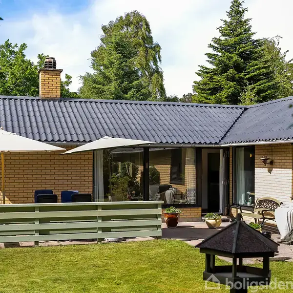 Hus med gult murværk og sort tag, roligt beliggende i en grøn have med terrasse. Parasol og havemøbler står på terrassen. Omkringliggende træer skaber en fredfyldt atmosfære.