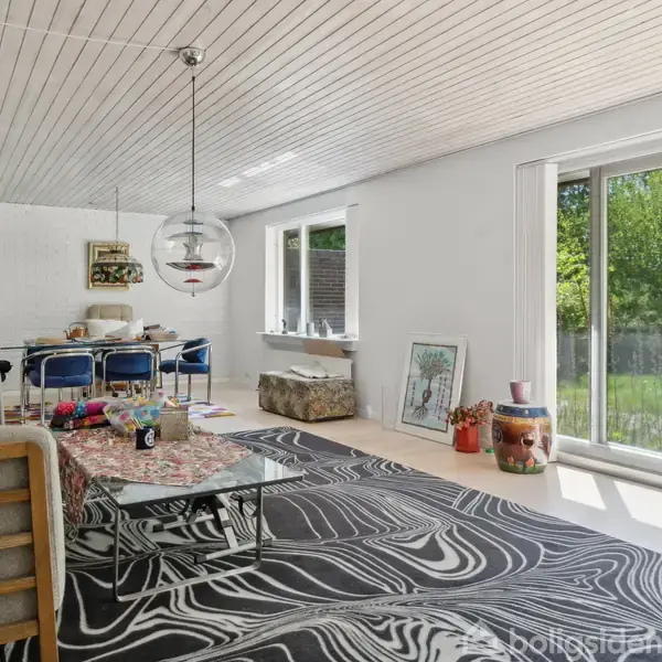 Et moderne stueområde indeholder en grå sofa og sofabord, placeret på et sort-hvidt mønstret tæppe. Store vinduer til højre viser en grøn have udenfor, og i baggrunden ses et spisebord med stole. En pendel hænger over bordet.