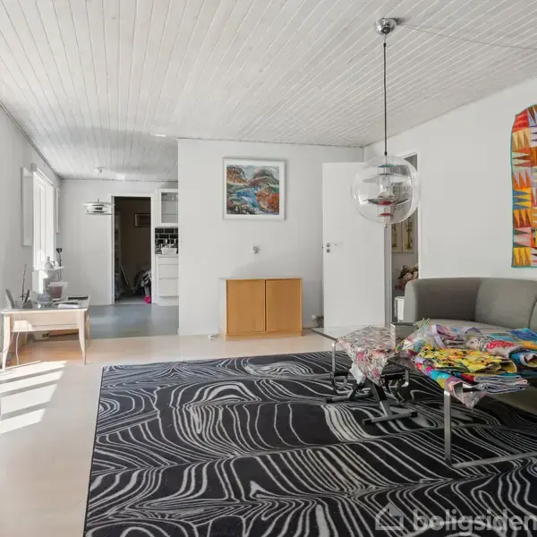 En moderne stue med en grå sofa, dekoreret med farverige puder og tæppe, står på et sort-hvidt mønstret tæppe. Væggene er hvide med et farverigt vægtæppe.