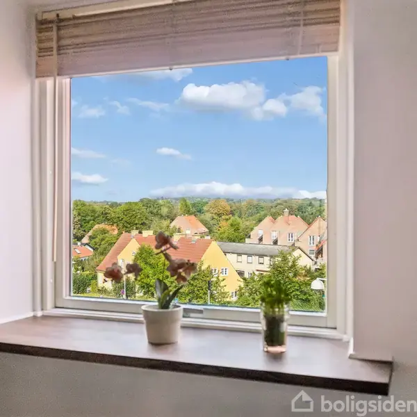 Et vindue med udsigt viser to blomsterpotter på vindueskarmen. I baggrunden ses gule huse med røde tage og grønne træer under en blå himmel med skyer.