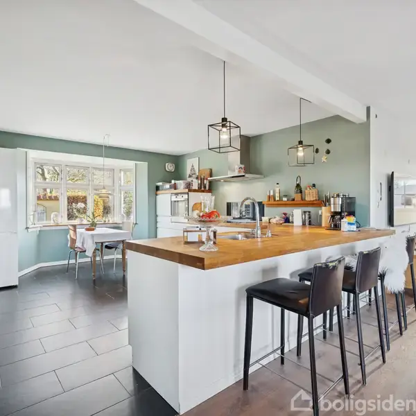 Køkken med en stor træbordplade og høje stole, moderne lamper over spisepladsen. Rummet er lyst med vinduer og spisestue i baggrunden. Grønlige vægge og flisegulv.