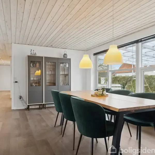 Et spisebord med grønne stole står i stuen. To gule lamper hænger over bordet. Store vinduer viser en terrasse udenfor i dagslys.
