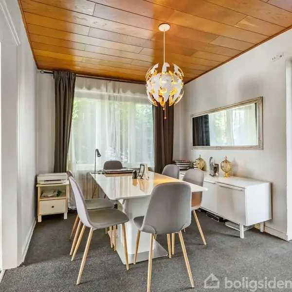 Spisebord med grå stole står i et rum med et hvidt skab og spejl på væggen, omkranset af et stort vindue med gardiner. En dekorativ loftlampe hænger over bordet.