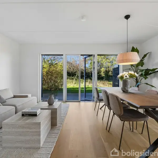 En moderne stue med en grå sofa og et sofabord, placeret mod et vindue med udsigt til en grøn have. Ved siden af står et spisebord med stole og en hængende lampe.
