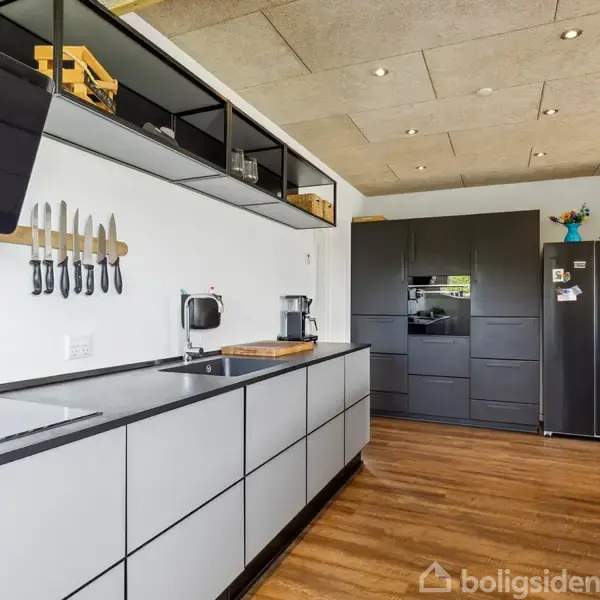 Sort og gråt køkken med knive på magnetisk holder, kaffemaskine ved siden af. Moderne køkkenskabe og træparketgulv danner rammen, og køleskabet har farverige magneter.