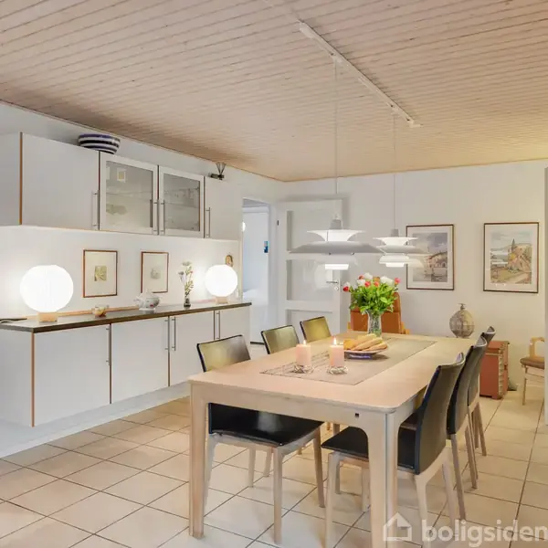 En spisestue med et langt træbord omgivet af seks sorte stole, dekoreret med blomster. Tre pendellamper hænger over bordet. Vægge prydes af kunst og skab, gulvet er flisebelagt.