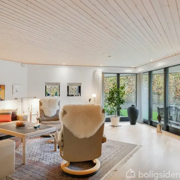 Stue med sofagruppe, bord og to stole med fåreskind, placeret på et tæppe. Store vinduer i baggrunden giver udsigt til en terrasse med planter. Væggene er dekoreret med kunst.