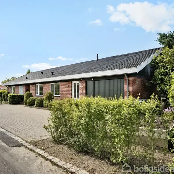 Et étplans murstenshus med sort tag står ved en asfalteret vej. Foran huset er der en stor belagt indkørsel og velplejede buske som hæk. Skyfri himmel i baggrunden.