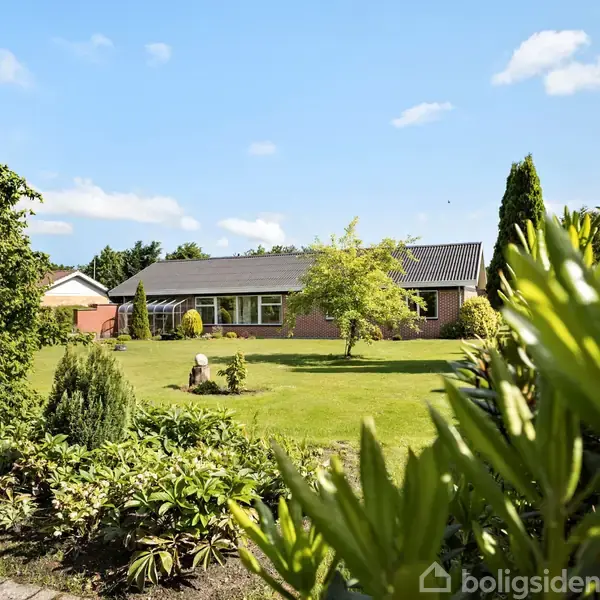 En enfamiliehus i mursten står midt i en velplejet have med grønne græsplæner og buske omkring. Solskin lyser scenen op under en blå himmel med spredte skyer.