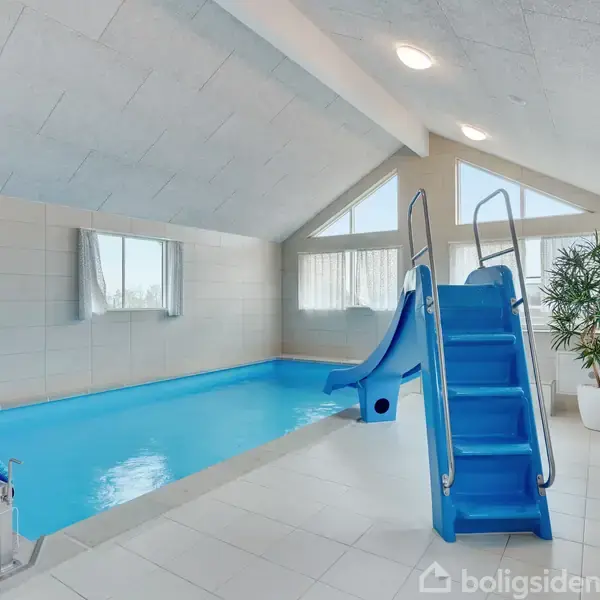 Indendørs swimmingpool med blå rutsjebane og rullende poolcover. Poolen er i et lyst rum med skrånende loft, store vinduer og en plante i hjørnet, der skaber en afslappende atmosfære.