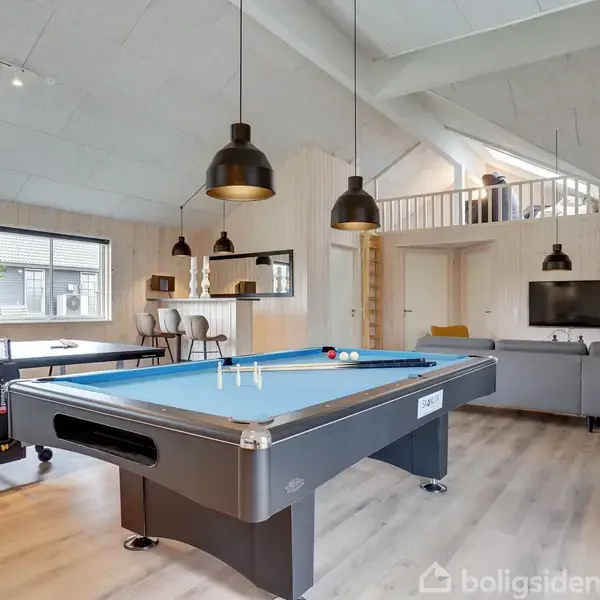 Poolbord står midt i et lyst rum ved siden af et bordtennisbord. Området har moderne møbler, en sofa, en tv-skærm, og flere hængelamper.