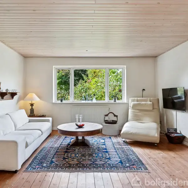 Stue med hvid sofa og lænestol foran et stort vindue. Et rundt træbord står på et orientalsk tæppe. Vægdør til højre og hylder med pynt til venstre.