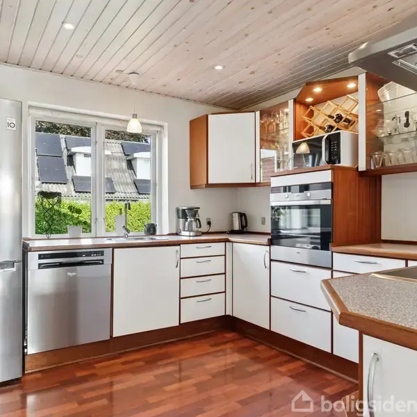 Et moderne køkken med hvide skabe og trædetaljer; et stort køleskab, indbygget ovn og opvaskemaskine. Gennem vinduet ses solpaneler på en tagbaggrund. Parkeret kaffemaskine og kedel på bordpladen.