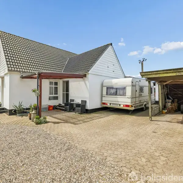 Hvidt hus står ved siden af en campingvogn under en carport. Grusområde og grønne marker omgiver stedet under en blå himmel.