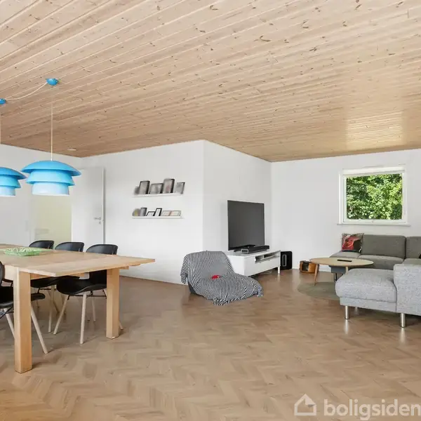 Spisestue med bord og seks stole under tre blå lamper, efterfulgt af stue med grå sofa, tv og reol. Trægulv og træloft skaber en hyggelig atmosfære.