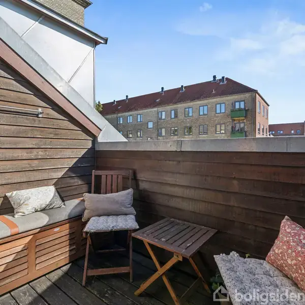 Lille altan med træbeklædte vægge og gulv, møbleret med en bænk, to stole og et lille bord. I baggrunden ses en række murstensbygninger under en blå himmel.