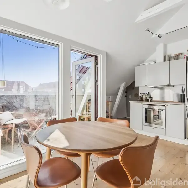Rundt spisebord med fire stole står i køkkenalrum ved siden af en stor, lys altandør. I baggrunden ses et moderne køkken med stål køleskab og hvide skabe.