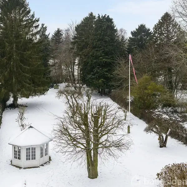 Et sneklædt haveområde med en pavillon og et flag på en høj flagstang. Store træer omkranser området, og en mindre bygning ses til venstre.