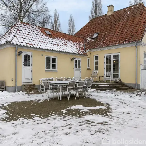 Et gult hus med rødt tegltag står i en snedækket haveomgivelser. Et stort udendørs bord med stole er placeret på en terrasse af fliser. Træer ses i baggrunden.