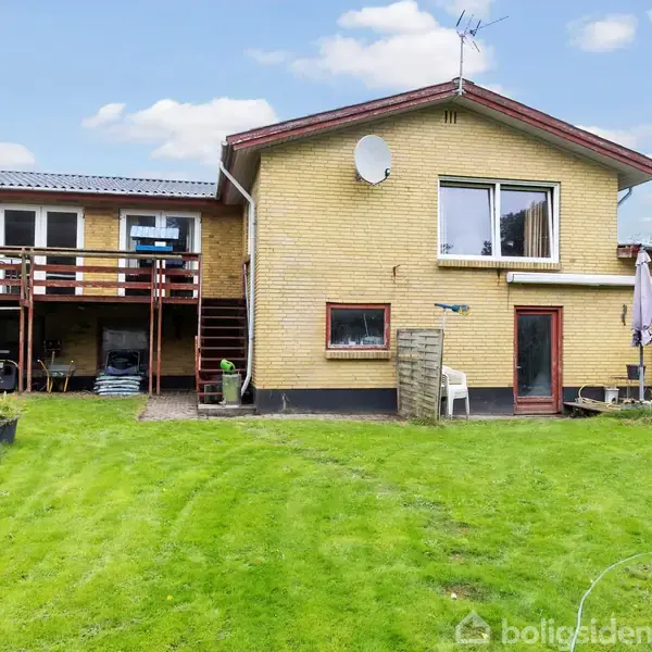 Murstenshus med et stort vindue og satellitparabol. Omkringliggende have har grønt græs, plantekasser og terrasse med bord og paraply. Klar blå himmel i baggrunden.