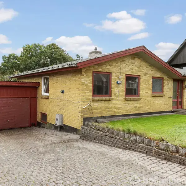 Et gulstenshus med rødt tag og tilhørende garage står på en brostensbelagt indkørsel. Græsplæne og buske omkranser huset under en blå himmel med få skyer.