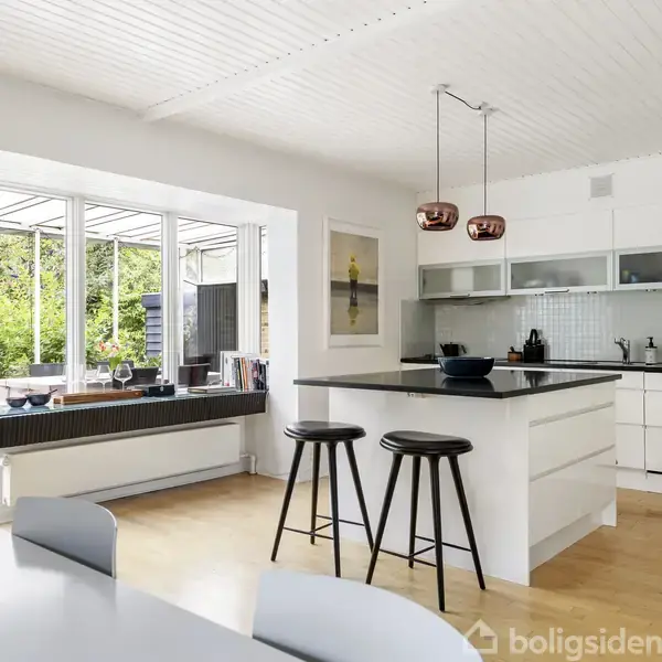 Køkken med hvidt interiør og sort køkkenbord, to barstole ved øen. Store vinduer med udsigt til frodig have. Moderne indretning med ophængte lamper og spisebord i forgrunden.