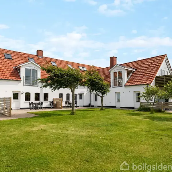 En stor, hvidbygget landejendom med rødt tegltag står i en velholdt græsplæne med træer. Huset har flere vinduer og altaner, der giver et landligt og rummeligt indtryk.