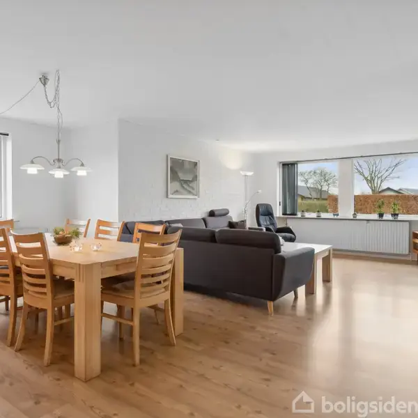 En åben stue med træbord og stole er midtpunktet, omgivet af sofa, store vinduer, og grønne planter. Lyse vægge og trægulv skaber en lys og indbydende atmosfære.