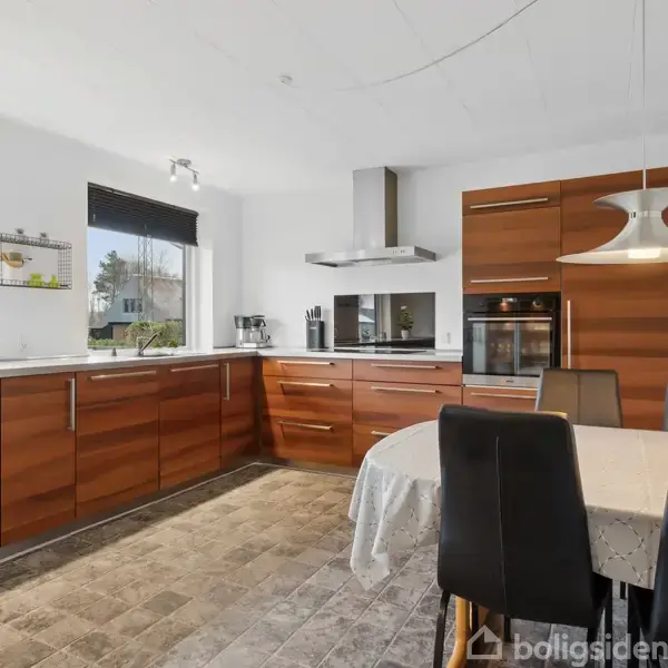 Køkken med træskabe, hvor der står et rundt bord med stole. Skabe indeholder indbygget ovn og emhætte. Store vinduer lader naturligt lys ind. Flisegulve tilføjer et rustikt præg.