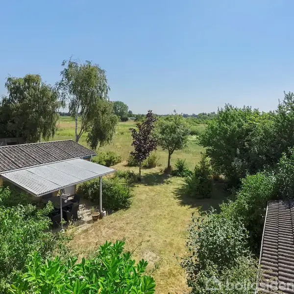 En baghave med græsplæne og træer omkranset af grønne buske. En overdækket terrasse på venstre side og en bygning med tegltag på højre side under en klar blå himmel.