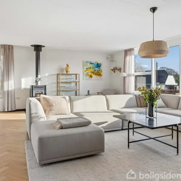 En stor, lys stue har en beige hjørnesofa, en sort pejs i hjørnet, og et stort vindue med udsigt til havet. Et sofabord står i midten med en vase.
