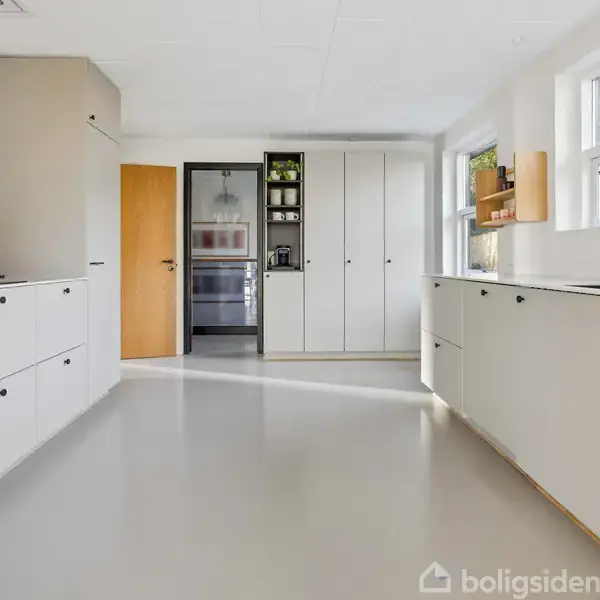 Et lyst køkken med hvide skabe og sorte greb. Skærebræt står på en skabsoverflade. Store vinduer til højre giver naturligt lys. I baggrunden ses en dør og indbygningsskab.