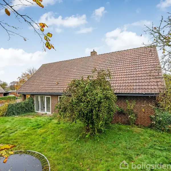 Et rødt murstenshus med et skråt tegltag står omgivet af en grøn have med buske og træer. En trampolin ses i forgrunden. Himmel med skyer i baggrunden.