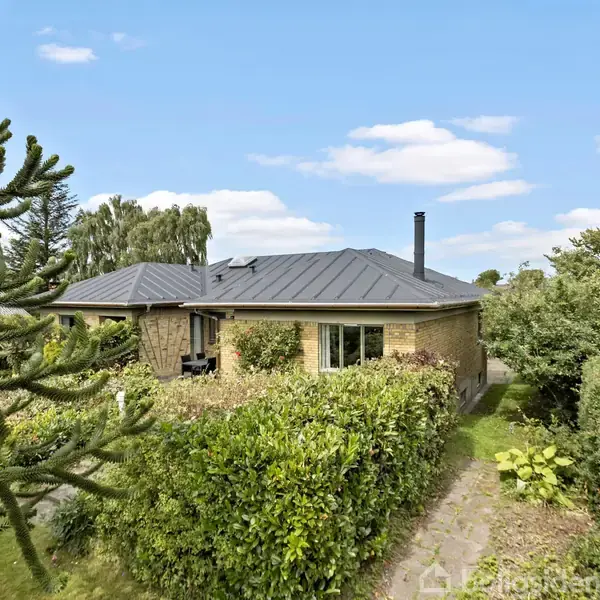 En etplans murstensvilla med sort tag placeret i en grøn have omgivet af buske og træer. Stien foran fører mod huset, og himlen er klar med lette skyer.