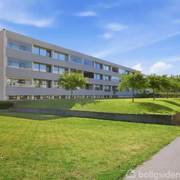 Et fleretagers lejlighedsbyggeri står i et grønt område med klippede græsplæner og små træer under en klar, blå himmel. Der er en sti langs bygningen.